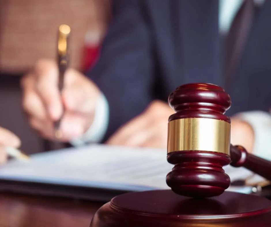 A wooden gavel with a gold band sits on a desk in the foreground, with a person writing on paper in the background.