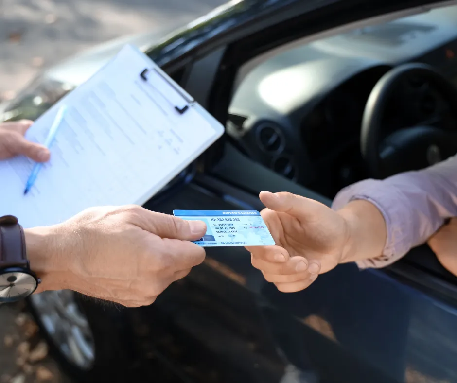 A person handing a driver's license to another person holding a clipboard with a pen next to a car.